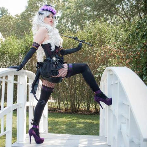 A model in a detailed gothic Lolita costume posing on a white arched bridge outdoors. The costume features a black corset with white ruffles, a black frilly skirt, fishnet stockings, and purple high heels.