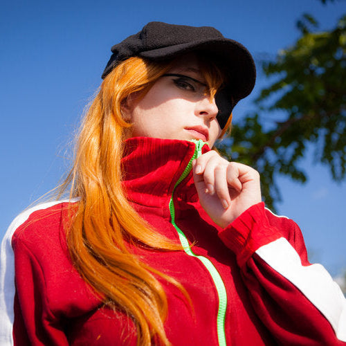 A woman outdoors wearing a red zip-up jacket with white side panels, a black cap, and long orange-red hair, set against a blue sky and blurred green trees.