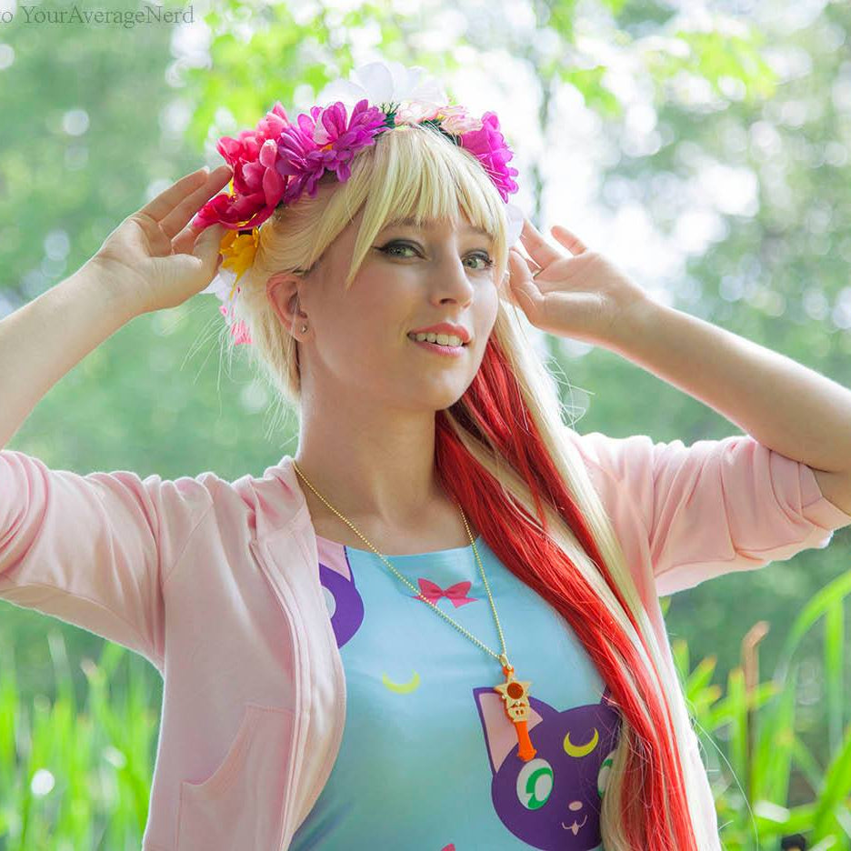 A woman outdoors wearing a pink flower crown, a light pink cardigan, and a light blue graphic T-shirt with a purple cat design. She has long blonde hair with red streaks and is posing with her hands near her head against a blurred green background.