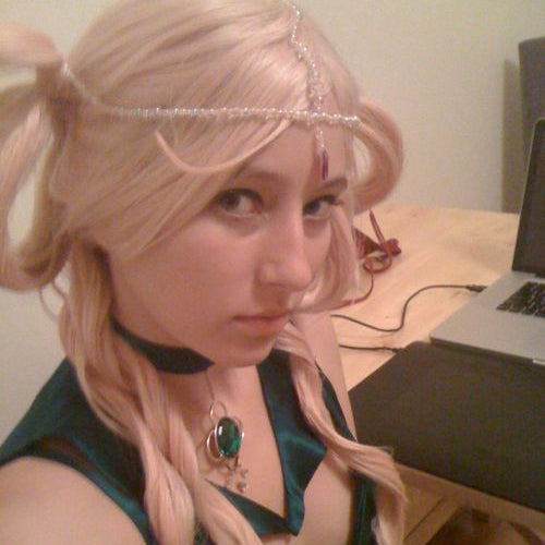 A woman wearing a light pink wig with a headpiece, a dark teal dress, and a green pendant necklace, photographed indoors near a desk and laptop.