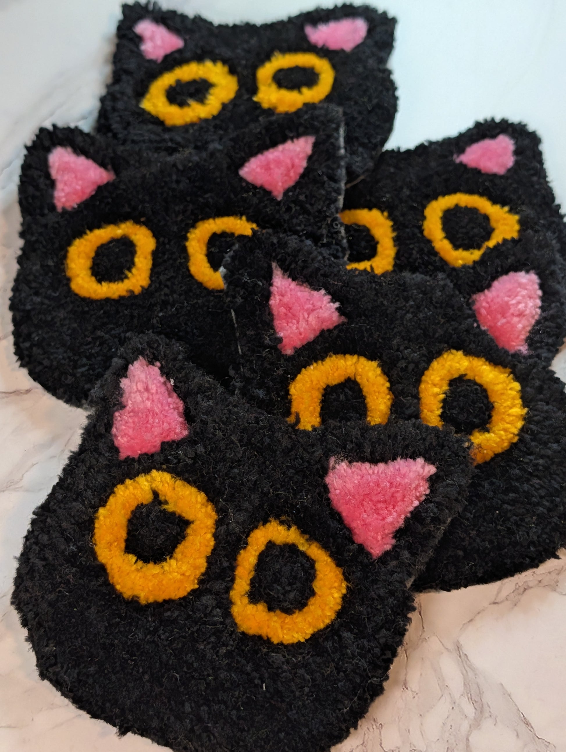 several Black cat-shaped tufted mug rug  with pink ears and yellow eyes on a marble background