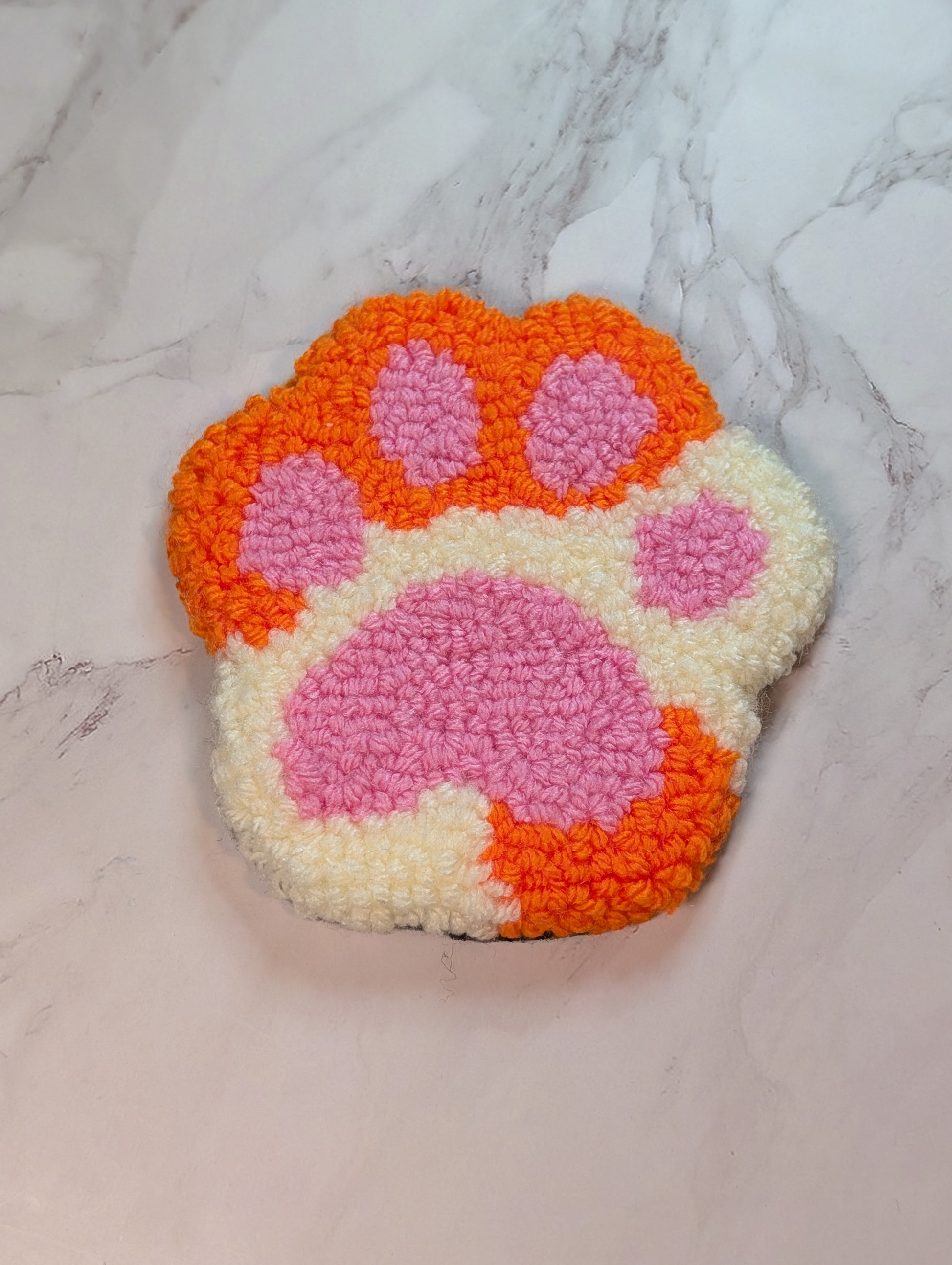 punch needle paw-shaped mug rug with orange and pink colors on a marble background