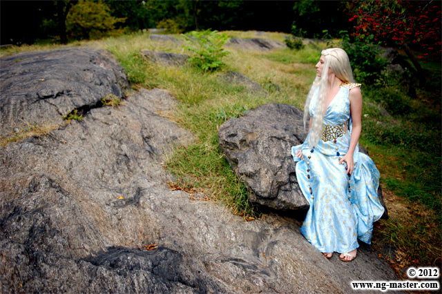 A woman with long platinum blonde hair sits on a large rock in an outdoor rocky landscape, wearing a flowing light blue gown with a metallic gold belt detail.