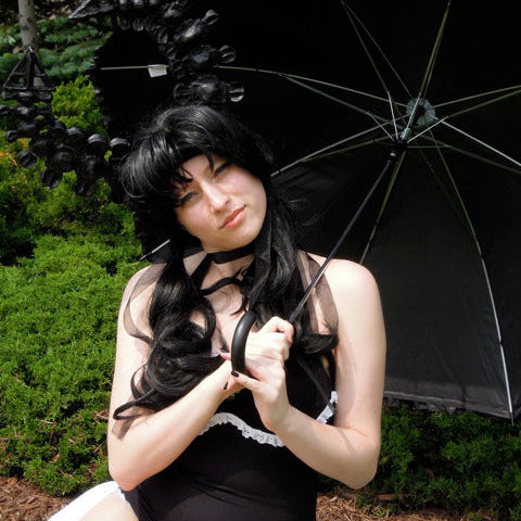A woman in a black gothic costume with a long black wig, holding a black umbrella outdoors on grass with bushes behind her.