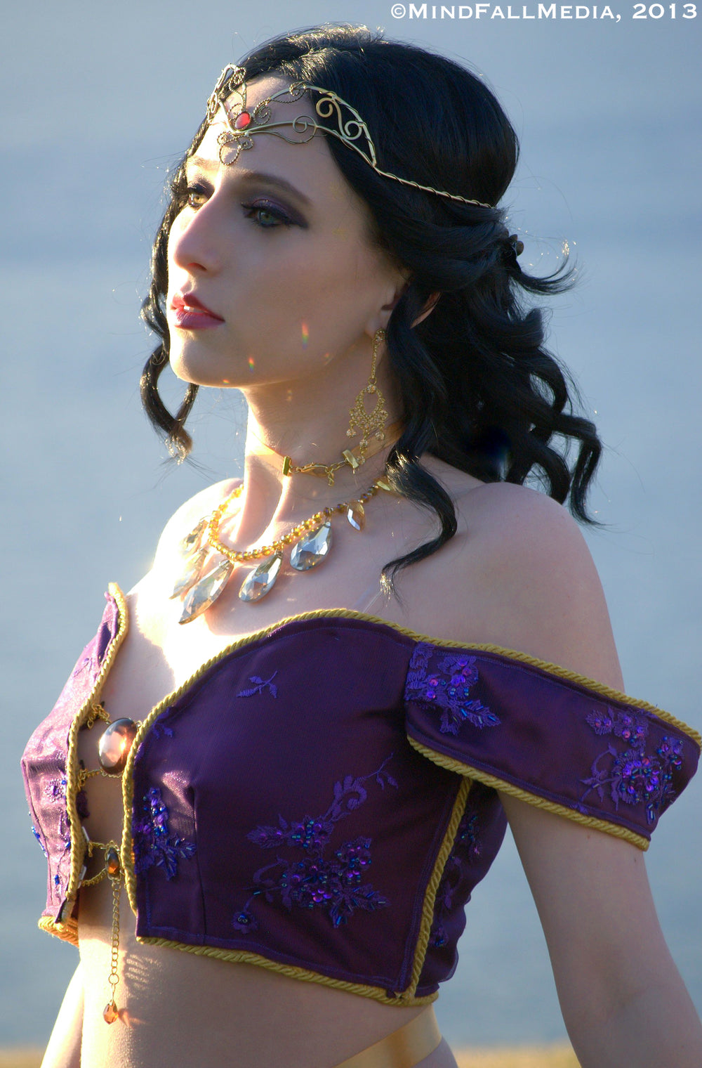 A woman wearing a purple and gold ornate off-the-shoulder crop top with floral embroidery, styled with gold jewelry and a headpiece against a soft blue background.