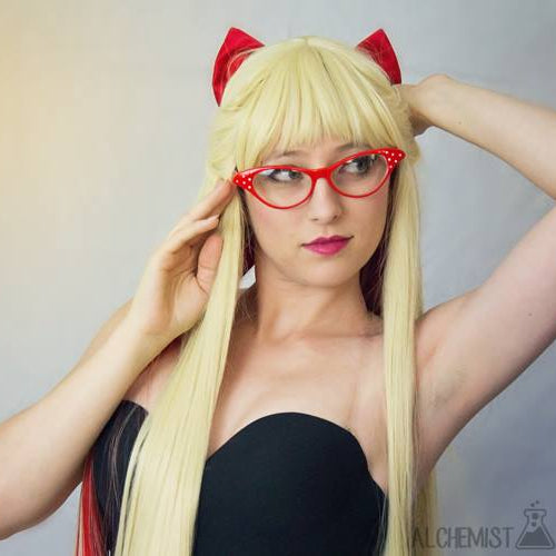 A woman posing in a blonde wig with red hair bow  and red cat-eye glasses, wearing a black strapless top against a gray studio background.