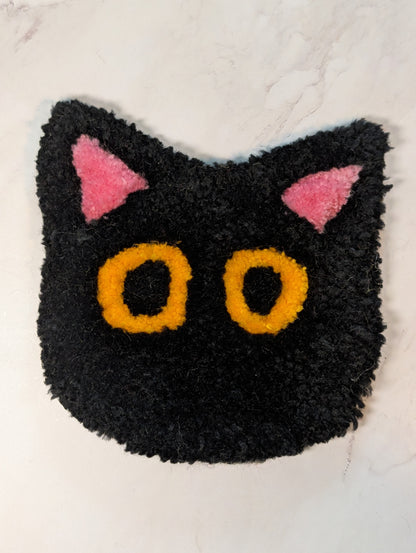 Black cat-shaped tufted mug rug  with pink ears and yellow eyes on a white background