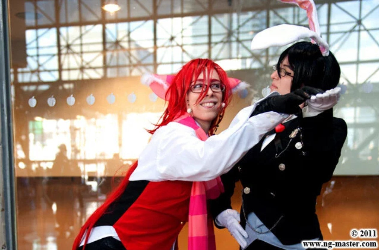 Two people in anime cosplay posing together in front of a glass window. The person on the left has bright red hair, glasses, and a white, red, and pink outfit with bunny ears.