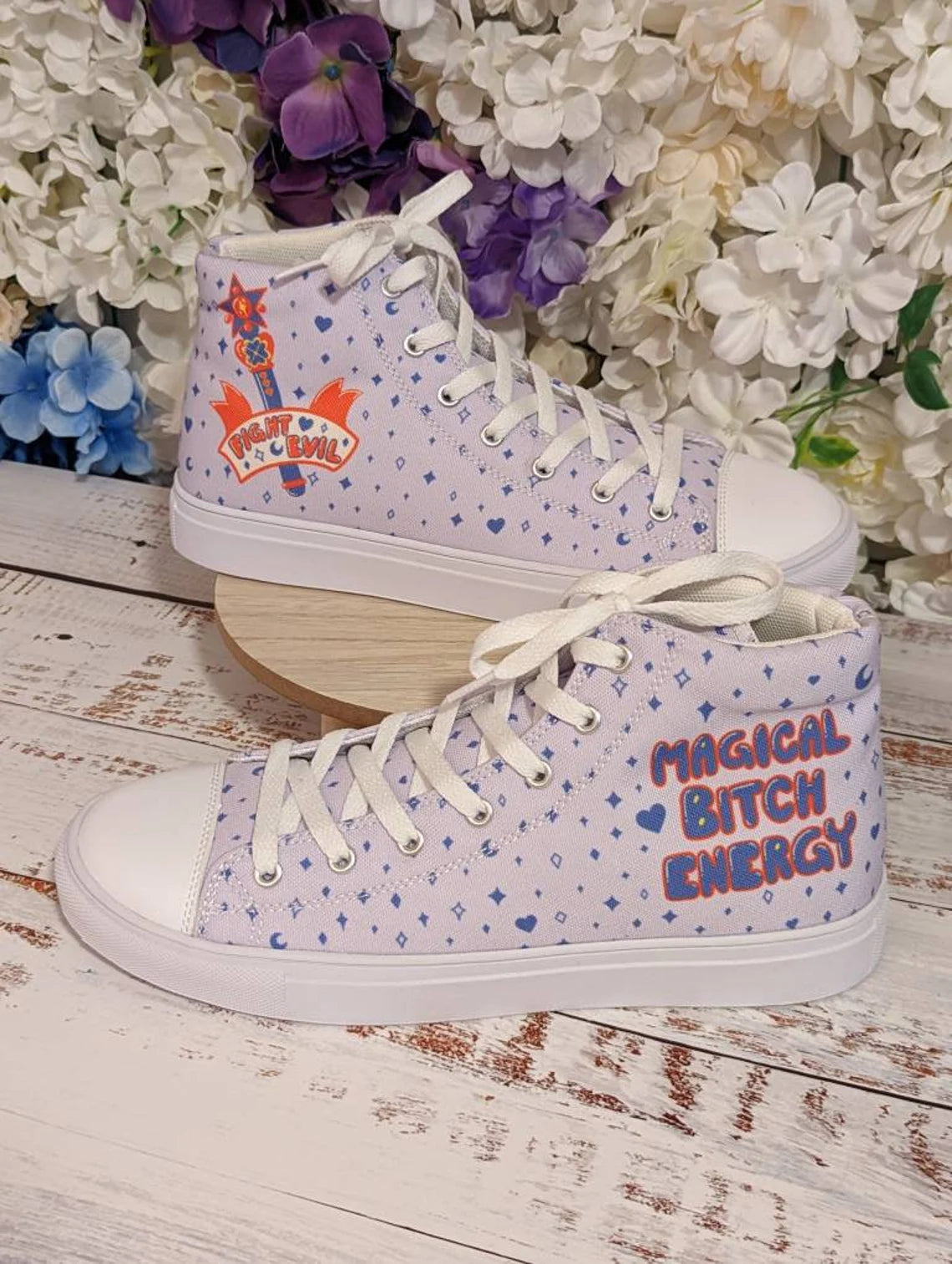 A pair of high top canvas shoes with a light purple background featuring a 'Magical Bitch Energy' statement and a magic transformation wand on one shoe, and a faux leather toe cap on the other. The shoes are positioned on a wooden surface with a floral arrangement and stars in the background.